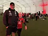 Flere personer står og sitter i et stort telt med grønt gulv mens en fotballkamp vises på storskjerm.