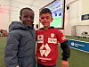To barn står foran en storskjerm i et lokale med fotballkamp på skjermen.
