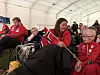 Flere personer i røde supporterklær sitter i et telt og ser på kamp på en skjerm.