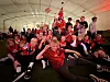 En stor gruppe mennesker i røde fotballtrøyer sitter og står samlet under et telt med norske flagg.