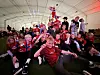 En gruppe mennesker i røde fotballdrakter sitter og står tett sammen i et telt med norske flagg.