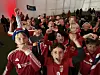 En gruppe mennesker i røde fotballdrakter jubler og vinker foran en storskjerm inne i et telt.
