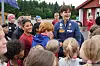 En gruppe barn samler seg rundt en smilende mann i blå jakke på en fotballbane.