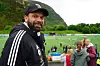 En smilende mann i svart Adidas-jakke står foran en fotballbane med barn og voksne i bakgrunnen.