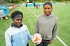 To jenter står på en fotballbane med en ball, mens flere barn trener i bakgrunnen.