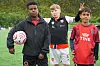 Tre gutter står på en fotballbane, to i drakter og én holder en fotball.
