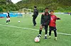 To barn står ved en fotball på en kunstgressbane, med mål, flere barn og en voksen i bakgrunnen.