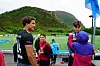 Barn og en trener står ved en fotballbane med fjell i bakgrunnen, mens en ung mann snakker med dem.
