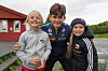 Tre barn smiler sammen med en fotballspiller utendørs ved en rød bygning.