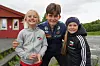 Tre barn smiler sammen med en fotballspiller utendørs ved en rød bygning.