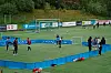 Barn og voksne er samlet på en fotballbane med små mål og blå presenninger langs sidelinjen.