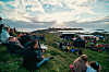Folk sitter på en grasbakke og ser utover et festivalområde med telt og scene ved sjøen.