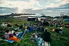 Folk sitter på tepper og stoler i en gressbakke med scene, telt og hav i bakgrunnen.