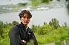 Ung mann i svart jakke står med armene i kors foran grønn natur og vann.