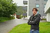 En ung mann står med armene i kors på en grønn plen ved en boligblokk.