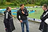 Tre menn står ved en fotballbane og snakker, med mål og reklameskilt i bakgrunnen.