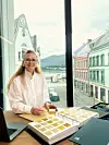 En smilende kvinne står ved et bord med en stor fargeprøvebok foran et vindu med utsikt mot by og fjord.