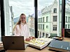 En kvinne sitter ved et skrivebord med laptop, prøver og dokumenter foran store vinduer.