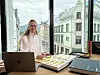 En kvinne sitter ved et skrivebord med laptop, prøver og dokumenter foran store vinduer.