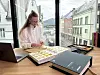 Person ved et skrivebord med laptop, mapper og fargeprøver foran et vindu med utsikt over bygårder.