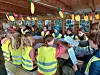 En gruppe barn sitter tett sammen innendørs og leser ark med en voksen i en gårdslignende bygning.