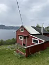 Rød hytte med hvitt tak ligger i skrått terreng ved fjorden under overskyet himmel.
