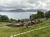 Rester av en utbrent hytte i grasbakke med fjord og fjell i bakgrunnen.