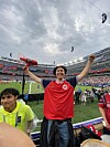 En person i rød drakt står på tribunen med armene hevet på et fotballstadion.