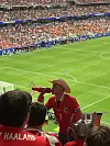 En mann i rød drakt og cowboyhatt står ved tribunene med en megafon, med fullsatt stadion og grønn bane i bakgrunnen.