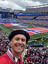 En smilende mann tar selfie foran et fullsatt stadion med store norske flagg på banen.