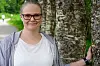 En kvinne med briller står ved en stor trestamme ved en sti i et grønt parkområde.