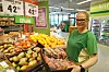 En smilende butikkansatt står ved frukt- og grøntdisken med kurver fulle av frukt og grønnsaker.