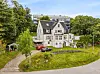 Stor hvit villa med mørkt tak, grønn hage og flere parkerte biler sett fra en skråning.