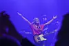 Gitarist med armene hevet på en scene med lilla lys og en musiker i bakgrunnen.