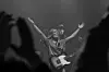 Musiker med gitar på scene, med armene i været, sett gjennom publikums hevede hender.