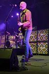 Gitarist synger og spiller på en scene med trommesett og mønstrede høyttalere i bakgrunnen.