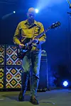 Gitarist spiller elgitar på en scene med blått lys og mønstrede forsterkere bak.