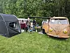 En gammel Volkswagen-buss, telt, campingstoler og bord står på gress i en grønn leirplass.