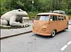 Oransje veteranbuss med takbagasje står på veien ved siden av en stor froskeskulptur.