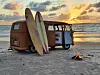 En vintagebuss på stranden med to surfebrett og et lite bål i forgrunnen.