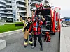 To brannfolk i dykkerutstyr står foran en rød brannbil og holder opp et norsk flagg.