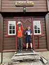 To barn står foran inngangen til en brun fjellhytte med røde detaljer og skiltet Reindalseter over døra.