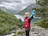 Et barn står på en stein med en blå leke, med fjell, dal og vann i bakgrunnen.