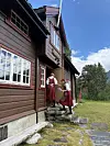 To personer i tradisjonelle klær står ved steintrappen utenfor en rødbrun trehytte med hvite vinduer.