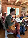 En mann spiller gitar inne i en tømmerhytte mens flere personer sitter og lytter.
