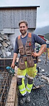 En arbeidsmann står på en byggeplass med verktøy i hånden, omgitt av armeringsjern, treverk og grus.