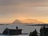 Solnedgang over hav og fjell med øyer i horisonten og bygninger i forgrunnen.