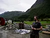 En mann står på et gårdstun med fjell, hus, paller og presenning i bakgrunnen.