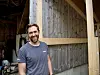 En smilende mann står foran en trelåve i landlige omgivelser.