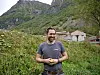 Mann står i grønt landskap foran gårdsbygninger og bratte fjell.